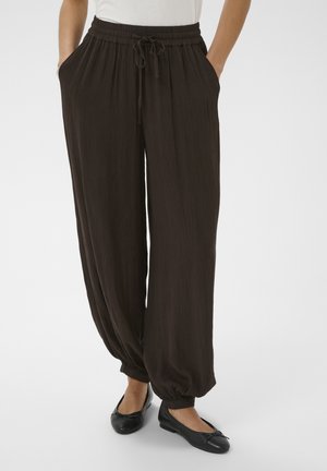 Kobieta ubrana w luźne ciemnobrązowe spodnie z elastycznymi mankietami i pasem, w czarnych balerinkach i białej bluzce, stojąca z rękami w kieszeniach.