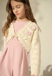 Cardigan in maglia crema con bordi a scallop e dettagli floreali in rosa, indossato su un vestito rosa a pieghe. Motivo testurizzato su tutta la superficie.