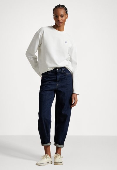 Camisa blanca de algodón de manga larga con un pequeño logotipo azul, combinada con vaqueros oscuros de tiro alto y zapatillas beige. Atuendo simple y casual.