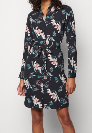 Vrouw draagt een zwarte, lange mouwen, knoopjurk met een roze en turquoise bloemenprint en een ceintuur, staand tegen een effen achtergrond.