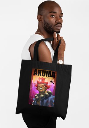 Homme avec barbe et montre tenant un sac fourre-tout noir avec Akuma de Street Fighter, aux cheveux rouges et grosses perles de prière.