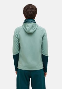 Man draagt een tweekleurige groene hoodie met de tekst "Peak Performance" op de kraag, kijkt weg tegen een effen lichte achtergrond.
