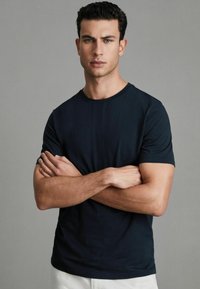 Tricou bleu marin cu guler rotund, fabricat din bumbac, cu mâneci scurte și o croială lejeră, purtat de un bărbat cu brațele încrucișate.