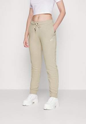 Personne portant un pantalon de survêtement beige Nike avec cordon de serrage et des chaussures blanches à plateforme lacées, posant avec une main dans la poche.