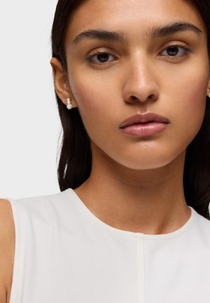 Femme aux cheveux foncés portant un haut blanc sans manches et des boucles d'oreilles en perles, présentée sur un fond clair uni.