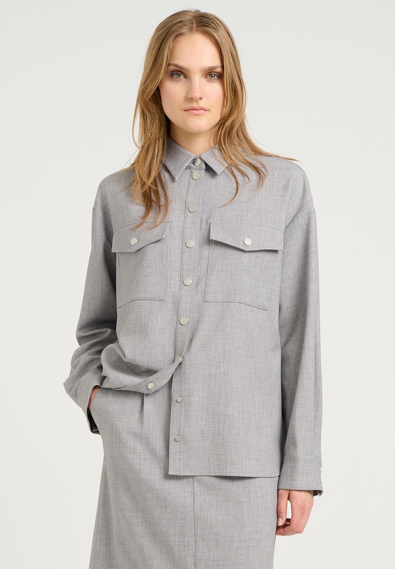 Camicia grigia con bottoni, maniche lunghe, due tasche sul petto e bottoni a pressione argentati. Presenta un tessuto morbido e strutturato con un design classico del colletto.