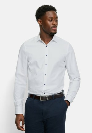 Weißes Langarmhemd mit Knöpfen und Besatz in Dunkelblau. Getragen in dunkelblauen strukturierten Hosen mit einem braunen Gürtel.