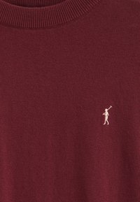 Burgundowa sweter wykonany z dzianiny z prążkowanym dekoltem i małym haftowanym logo w kremowym kolorze na lewej piersi. Gładka tekstura.