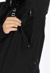 Giacca sportiva nera con tasca con zip, texture liscia e dettagli hardware decorativi. Etichetta del marchio visibile all'interno della zip.