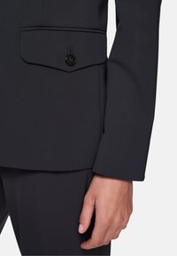 Dark gray suit jacket featuring a structured design, textured fabric, a chest pocket with a black button, and fitted sleeves.