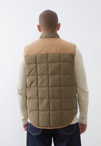 Uomo che indossa un gilet trapuntato bicolore marrone sopra una camicia a maniche lunghe crema, in piedi rivolto verso un muro bianco semplice.