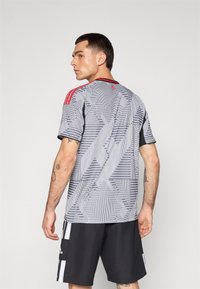 Maglia atletica grigia con un motivo geometrico, dettagli rossi sulle spalle, maniche corte e colletto. Abbinata a pantaloni corti neri.