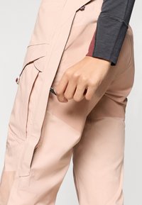 Calças em rosa pálido, impermeáveis, com bolsos laterais e um design elegante. Apresentam uma textura suave e costuras de destaque em tons mais escuros.