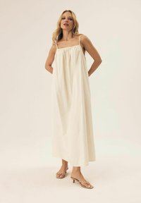 Cremfarbendes Maxikleid mit dünnen Trägern, gerafftem Oberteil und lässiger Silhouette. Der Stoff wirkt leicht und glatt.