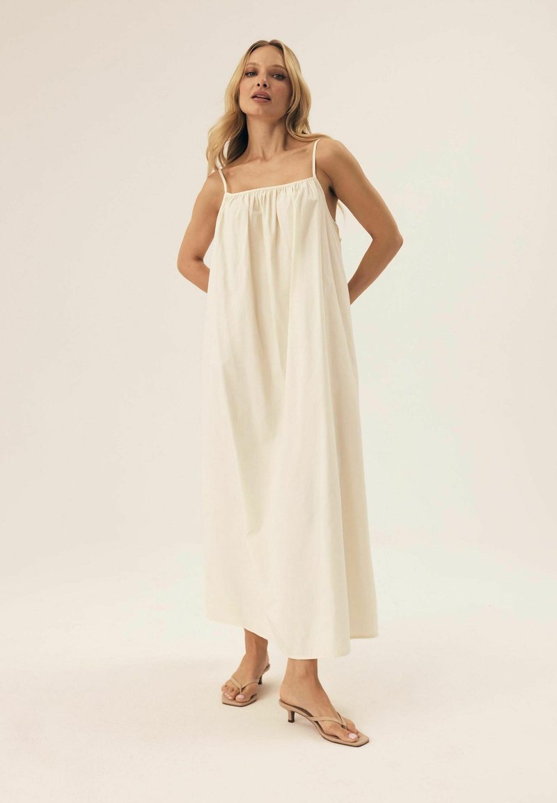 Cremfarbendes Maxikleid mit dünnen Trägern, gerafftem Oberteil und lässiger Silhouette. Der Stoff wirkt leicht und glatt.