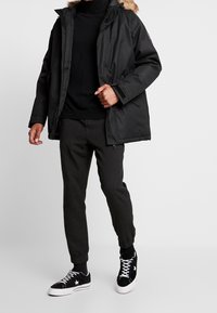 Mustat parka, jossa on turkiskuvioitu huppu, vetoketjukiinnitys ja sivutaskut; yhdistetty tummanharmaisiin housuihin ja mustiin lenkkareihin, joissa on valkoinen tähtikuvio.