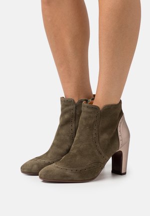 Botas de ante verde oliva hasta el tobillo con punta afilada, con detalles de brogue y un tacón de bloque metálico color beis. Textura suave y diseño elegante.