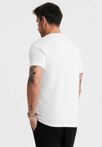 Bílá bavlněná tričko s krátkými rukávy a kulatým výstřihem, z pohledu zezadu, s viditelnými tetováními na levé paži.