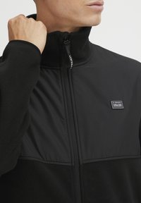 Mand iført sort zip-up jakke med fleeceærmer og bryst, holder lynlåsen tæt ved den høje krave med synlig logo-patch på brystet.