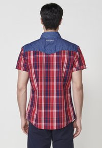 Kurzarmhemd mit einem rot-blauen Karomuster, einem Jeans-Ausschnitt und einem abgerundeten Saum. Eine gestickte Detail auf der Rückseite ist in der Nähe des Kragens sichtbar.