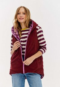 Kobieta ubrana w bordowy kamizelkę z korduroyu z kwiatową podszewką, nałożoną na fioletową i białą, w paski, długą koszulę oraz niebieskie dżinsy, poprawia zamek w kamizelce.