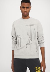 Grå sweatshirt med ett texturerat grafiskt mönster i svart och gult. Ribbstickade muddar och fåll, tillverkad av mjukt, lätt material.
