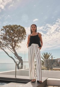 Haut sans manches noir avec des bords volantés, associé à un pantalon large rayé blanc. Cadre en plein air avec un arbre et vue sur l'océan.