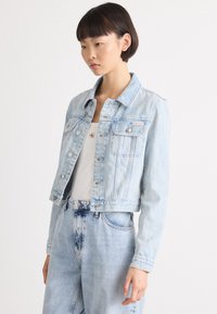 Lys blå denimjakke med en kortere design, frontlommer og sølvfarget detaljer. Har klassisk krage og knappeåpning.