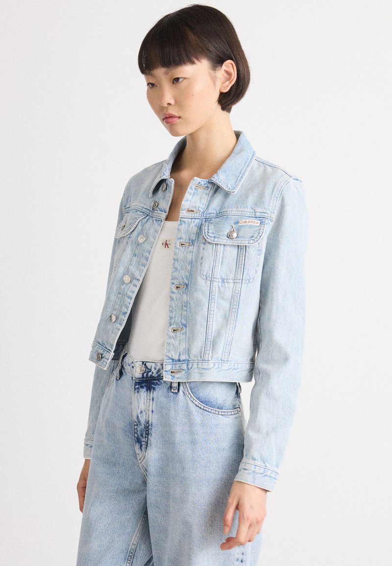 Lys blå denimjakke med en kortere design, frontlommer og sølvfarget detaljer. Har klassisk krage og knappeåpning.