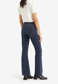 Jeans acampanados de lavado oscuro con cintura alta, que presentan bolsillos traseros y una etiqueta de marca sutil, combinados con una blusa de color claro y botas de tobillo.