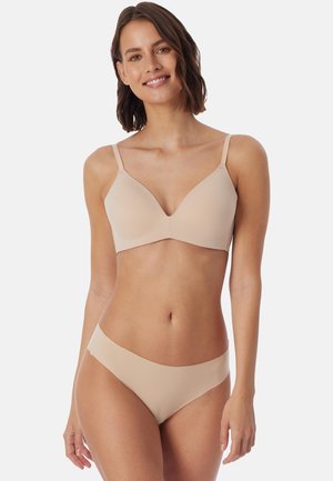 Frau mit kurzen braunen Haaren lächelt, trägt passenden beigen BH und Unterwäsche vor einem schlichten weißen Hintergrund.