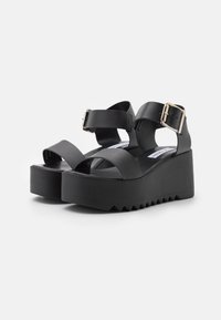 Sandalias de plataforma negras con dos correas anchas, detalle de hebilla metálica, suela de goma texturizada y una cuña gruesa para mayor altura.