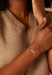 Zilveren armband met het woord "MOM" prominent weergegeven. Draagt op een gebruinde pols, tegen een gestructureerde beige gebreide trui.