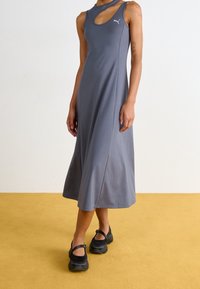 Robe grise sans manches avec un détail asymétrique découpé, en tissu lisse. Portée avec des chaussures noires, debout sur un sol jaune.