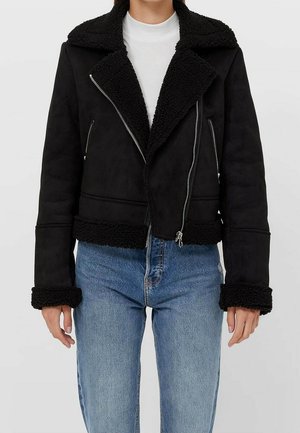 Veste courte en shearling noir avec col large et fermetures éclair argentées portée sur un top blanc et un jean bleu.