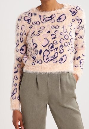 Pull court en tissu moumoute avec imprimé léopard rose et violet, associé à un pantalon taille haute vert olive, main posée dans la poche.