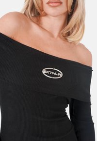 Schwarzes, geripptes Off-Shoulder-Top mit einem silbernen ovalen Logo-Akzent in der Mitte. Der Stoff wirkt glatt und hat eine figurbetonte Silhouette.