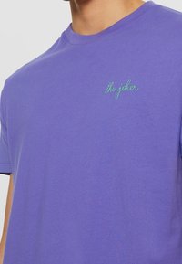 Lila t-shirt av bomull med rund hals och korta ärmar, med grön broderad text "the joker" på framsidan.