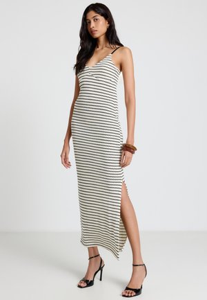 Frau trägt ein ärmelloses, schwarz-weiß gestreiftes Midikleid mit seitlichem Schlitz, schwarze High-Heel-Sandalen und braune Armbänder am Handgelenk.