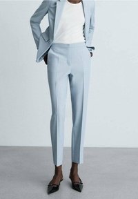 Persoon in een lichtblauw op maat gemaakt pak met cropped broek en zwarte spitse slingback hakken voor een effen achtergrond.