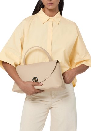 Žena v světle žluté košili a krémových kalhotách drží béžovou koženou kabelku s zakřivenou klopou a zlatým zapínáním.