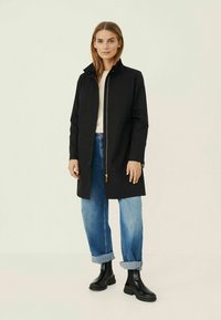 Black waterproof coat with a high collar, front zipper, and side pockets, paired with rolled-up blue denim jeans and black ankle boots.