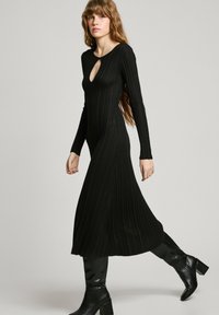 Black ribbed knit dress with long sleeves, keyhole neckline, and flared hem. Worn with knee-high black boots, showcasing a fitted silhouette.