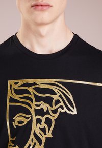 Camiseta negra con cuello redondo que presenta un contorno dorado brillante de un diseño de rostro clásico. La tela parece suave con una textura lisa.