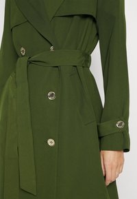 Gros plan d'un manteau vert olive à manches longues avec ceinture, boutons sur le devant, une poche et une lanière de manchette boutonnée, porté sur le torse d'une personne.