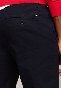Person som har på seg marineblå bukser med en baklomme med knapp og en liten rødt, hvitt og blått logo, sammen med en rød genser.