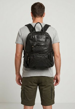 Mand iført grå t-shirt og olivengrøn cargo shorts med en sort læderrygsæk mærket "VENICE" på en lys baggrund.