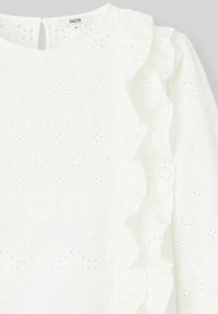 Blouse blanche avec motif floral en œillet, coutures latérales froncées et fermeture en goutte d'eau à l'arrière. Texture douce et tissu léger.