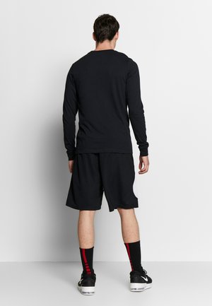 Maglietta nera a maniche lunghe e pantaloni corti coordinati, entrambi realizzati in un materiale liscio. I pantaloni corti hanno tasche laterali e dettagli bianchi. Sneakers nere con dettagli rossi.