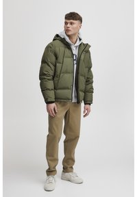 Green puffer jacket with a hood, zip closure, and ribbed cuffs, layered over a gray sweatshirt and paired with tan pants and white sneakers.